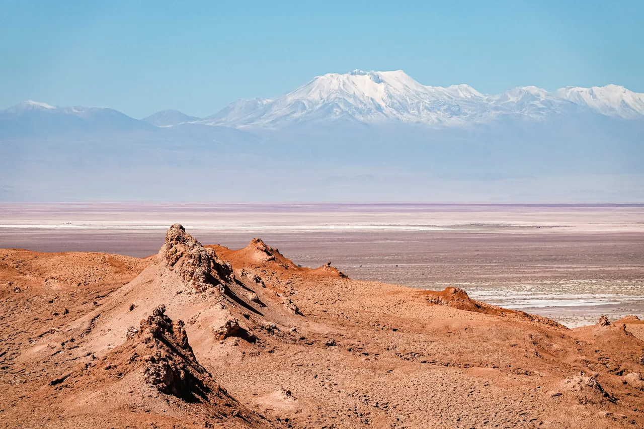 Formations rocheuses ocre du désert d’Atacama, avec une vaste étendue saline et les sommets enneigés de la cordillère des Andes en arrière-plan.